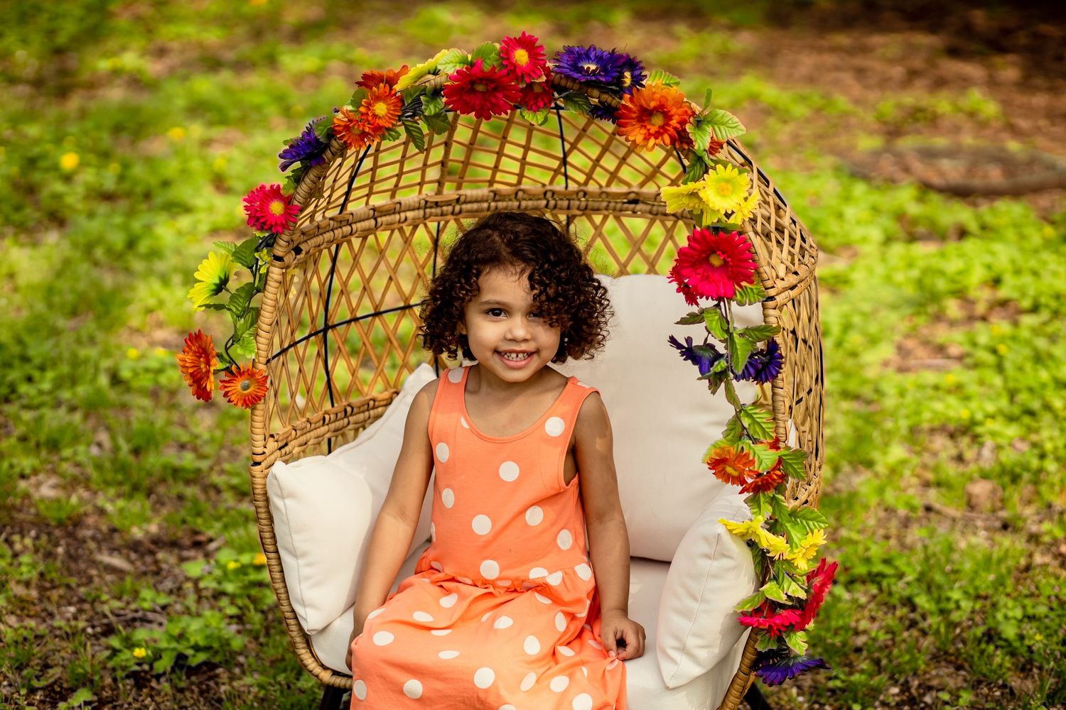 Young girl in orange polka dot dress sitting in floral wicker egg chair outdoors.