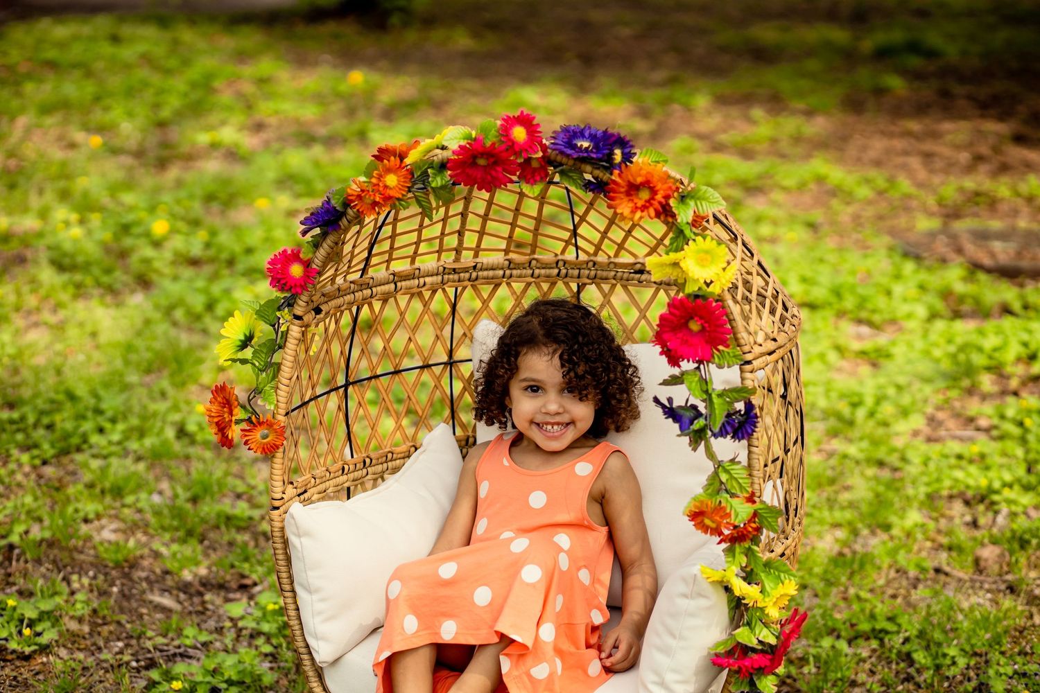 A smiling young girl in an orange polka dot dress sits in a floral wicker chair outdoors.