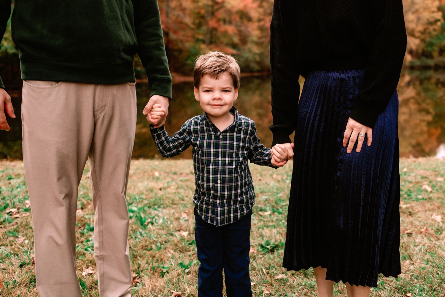 A young boy in a plaid shirt smiles while holding hands with his parents in an autumn outdoor setting.