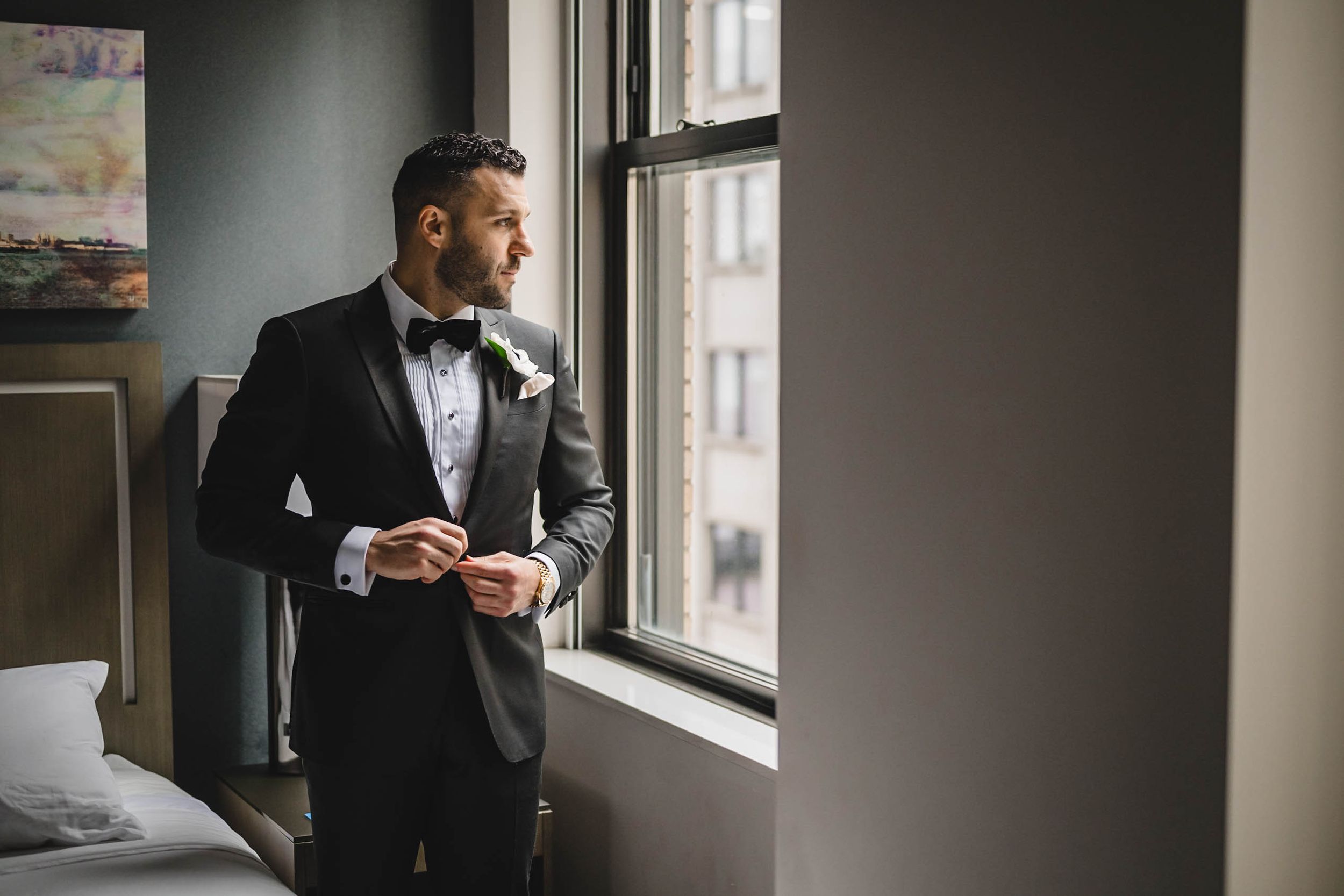 A groom in formal attire gazes out a window while holding a drink in a dimly lit room.