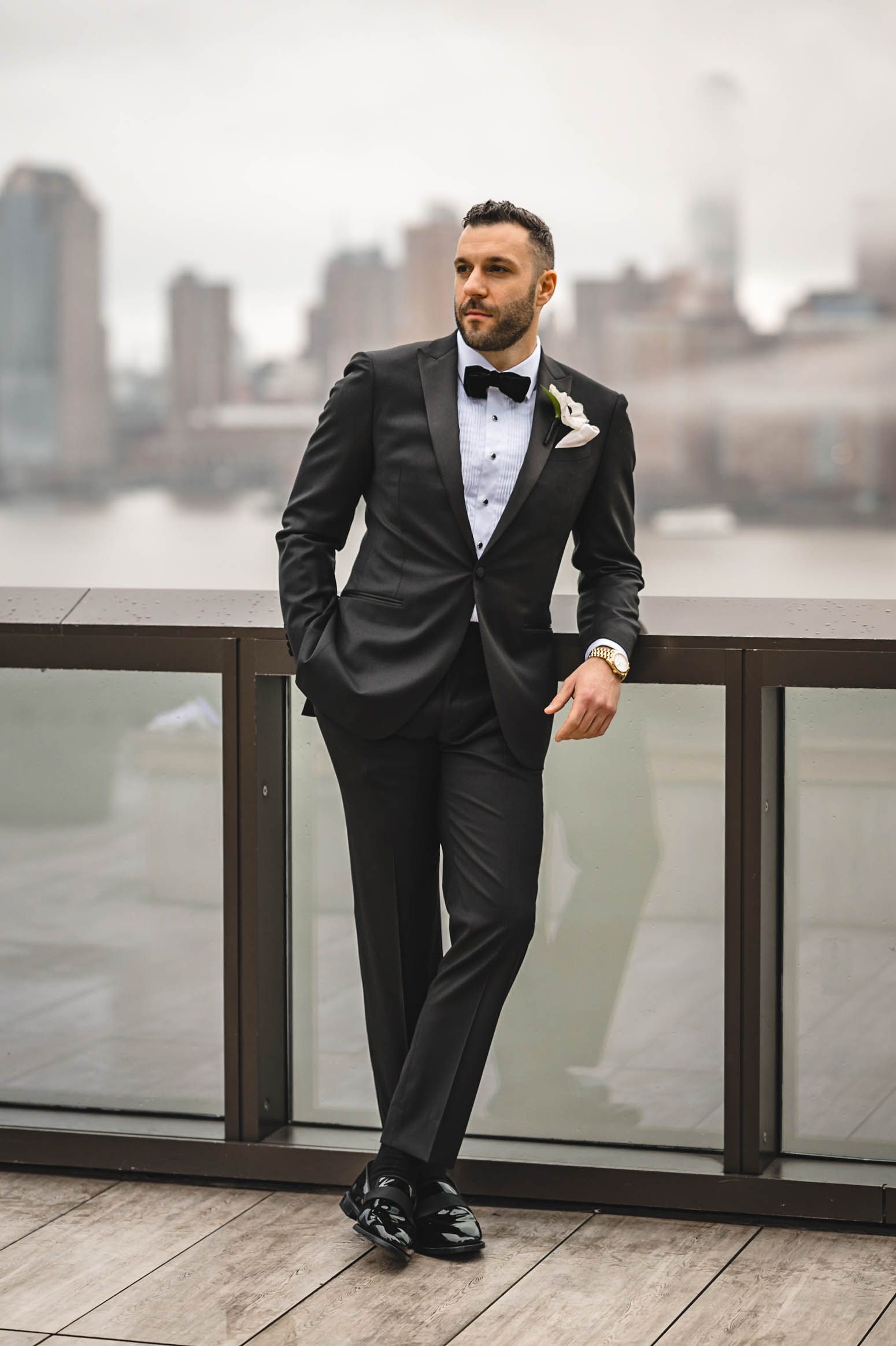 A formal portrait of a groom in tuxedo standing against city skyline backdrop on a deck.