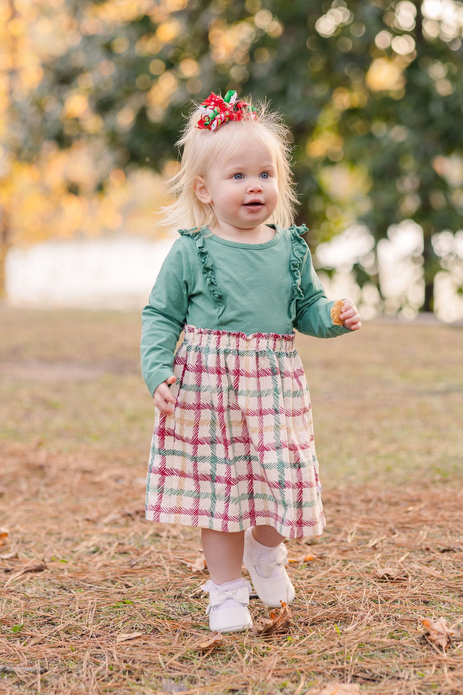 Toddler in green top and plaid dress walking outdoors with festive hair bow.