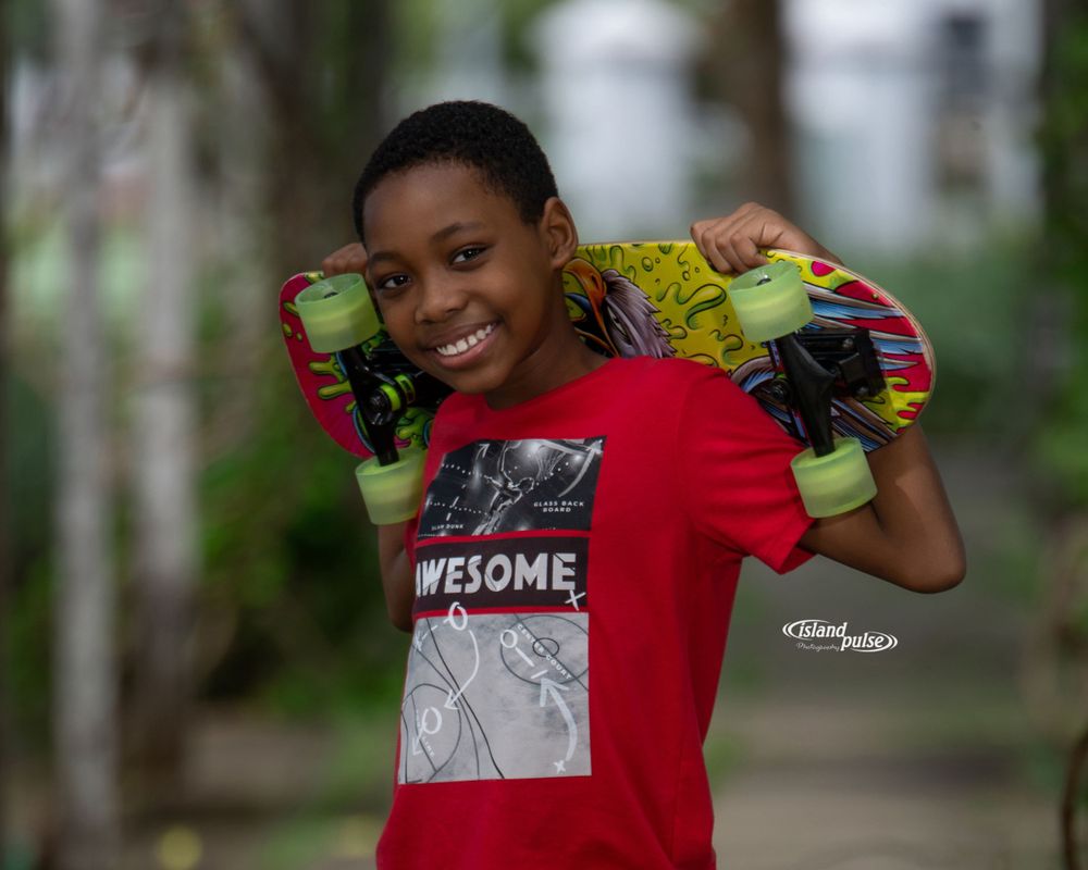 Smiling young boy in a red AWESOME t-shirt holding a red and yellow skateboard on his shoulders outdoors.