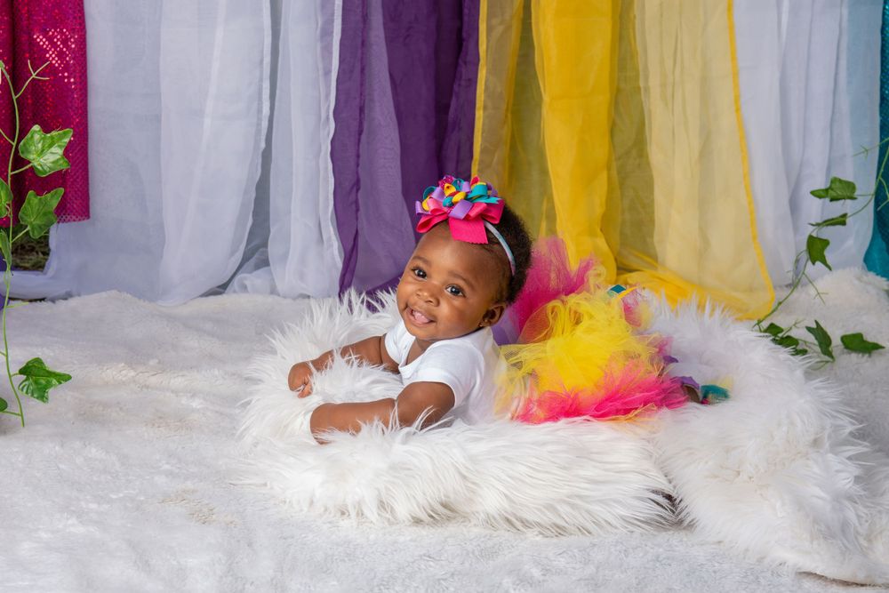 Smiling baby girl in colorful tutu and bow headband lying on white fur rug with rainbow fabric backdrop.