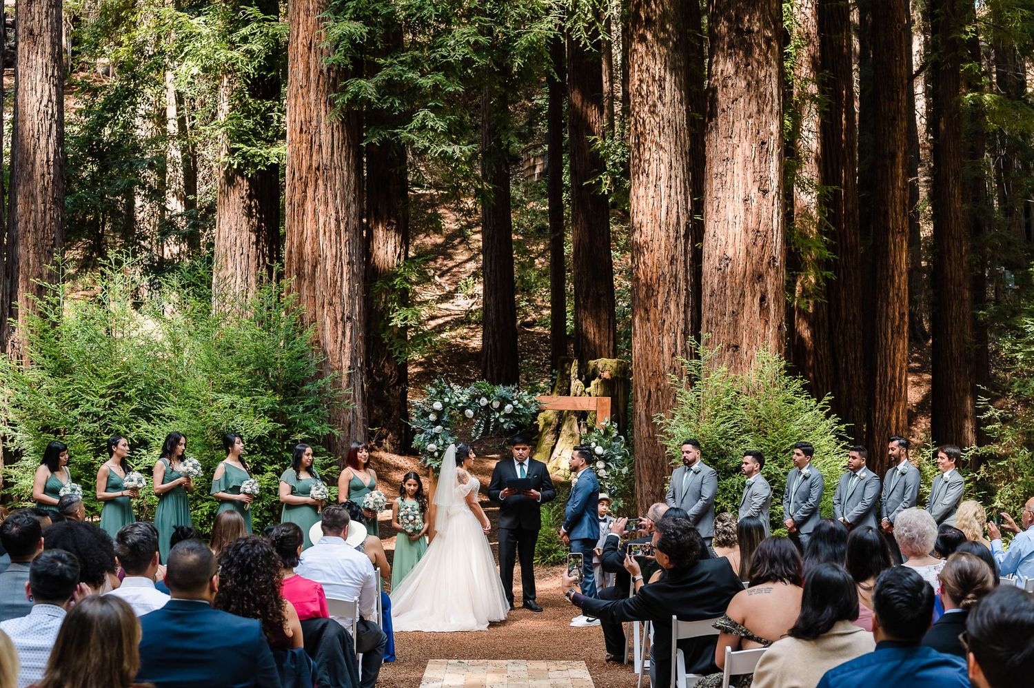 An outdoor forest wedding ceremony surrounded by towering redwood trees and seated guests.