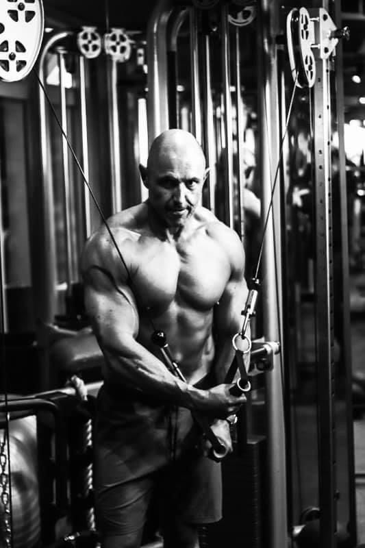 Bald muscular man performing cable crossover chest exercise on gym machine shirtless.