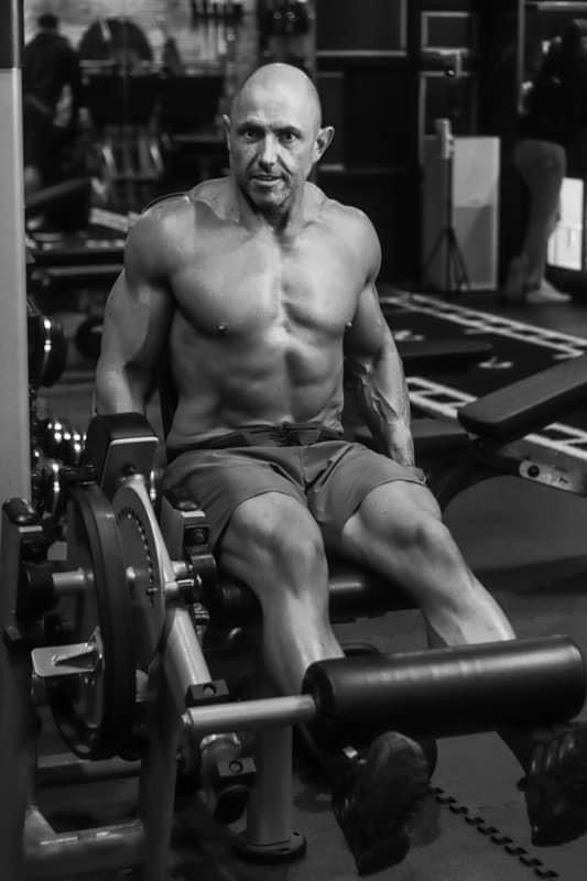 Bald shirtless bodybuilder resting on leg press machine in a gym between sets.