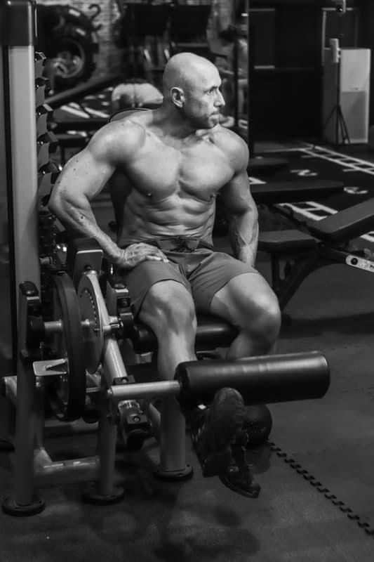 Shirtless bald bodybuilder with tattoos sitting on leg press machine in gym.