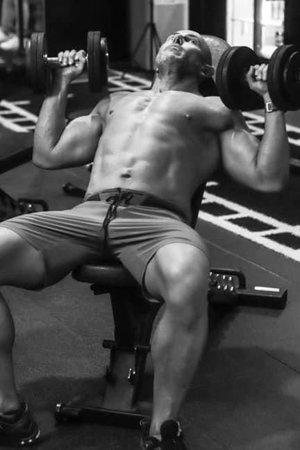 Muscular shirtless man performing dumbbell incline bench press exercise in gym.