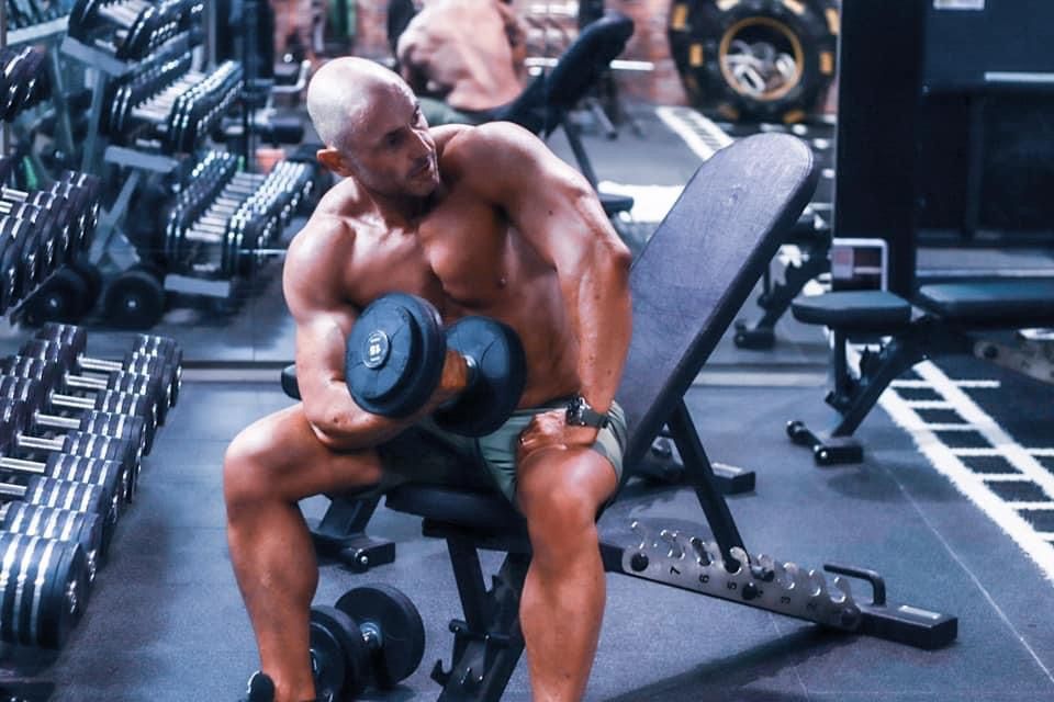 Muscular bald man performing concentration dumbbell bicep curls on bench in gym.