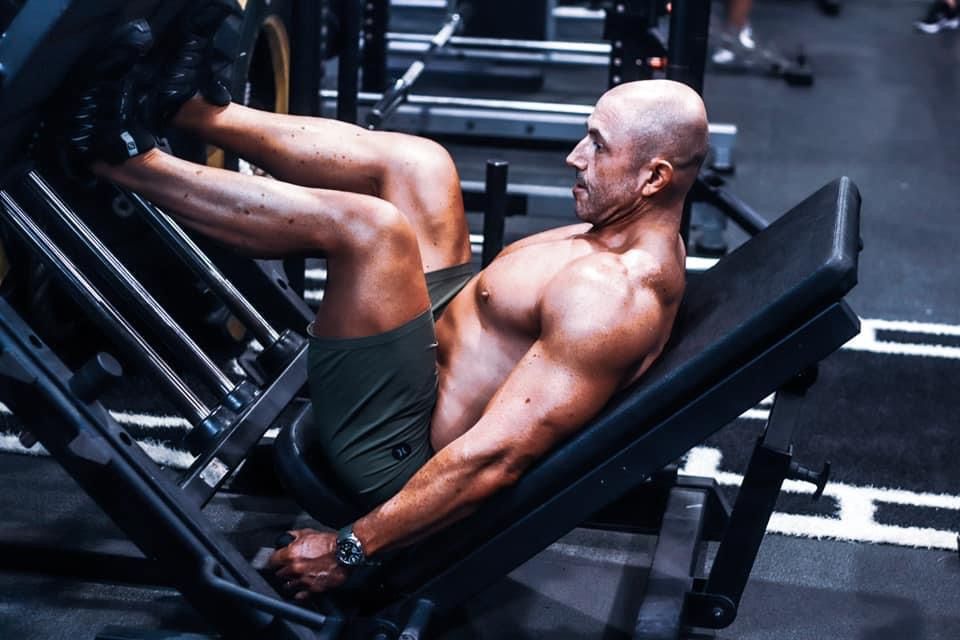 Bald shirtless bodybuilder performing leg press exercise on machine in gym.