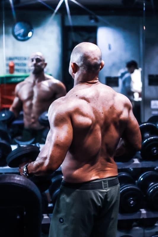 Bald muscular bodybuilder displaying back muscles while facing gym mirror with dumbbells.