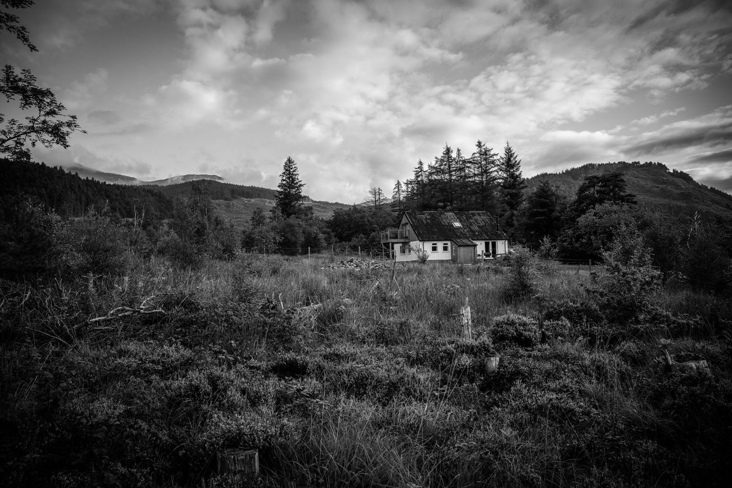 Monochrome landscape of a remote cottage nestled among hills with dramatic cloudy sky.