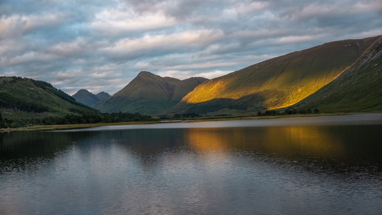 Tranquil mountain lake landscape with sunlight hitting peaks and dramatic cloudy sky reflections.