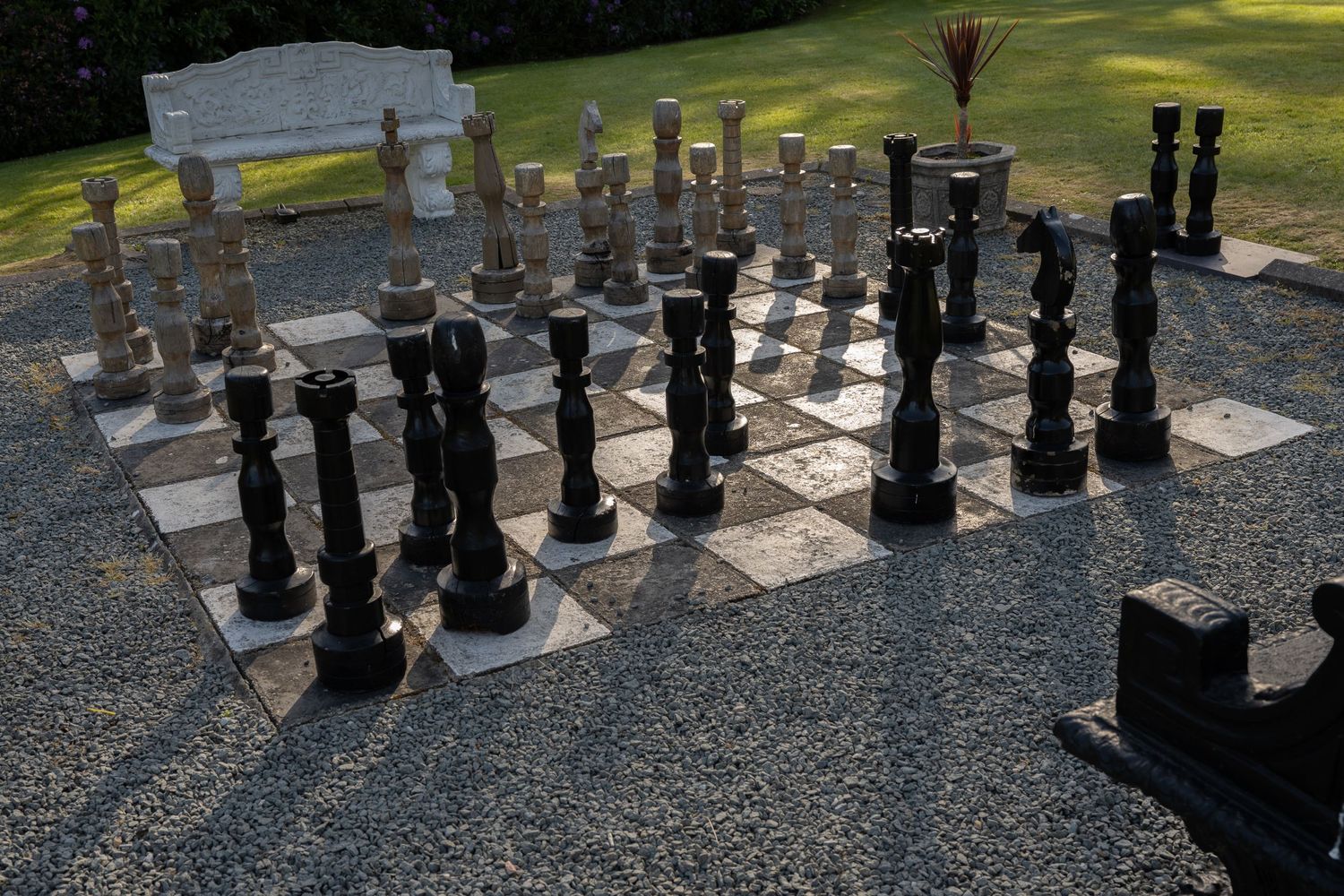 Large stone and wood outdoor chess set with carved pieces displayed on a patterned board near a bench.