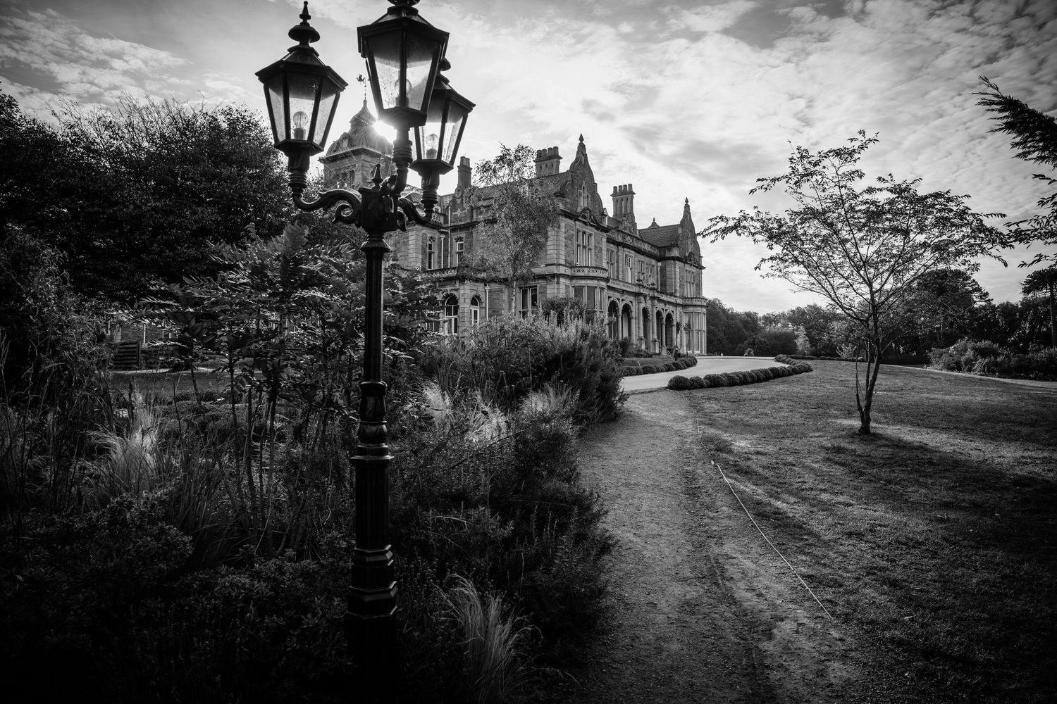 Black and white image of a Victorian mansion with ornate lamp posts along a curved pathway.