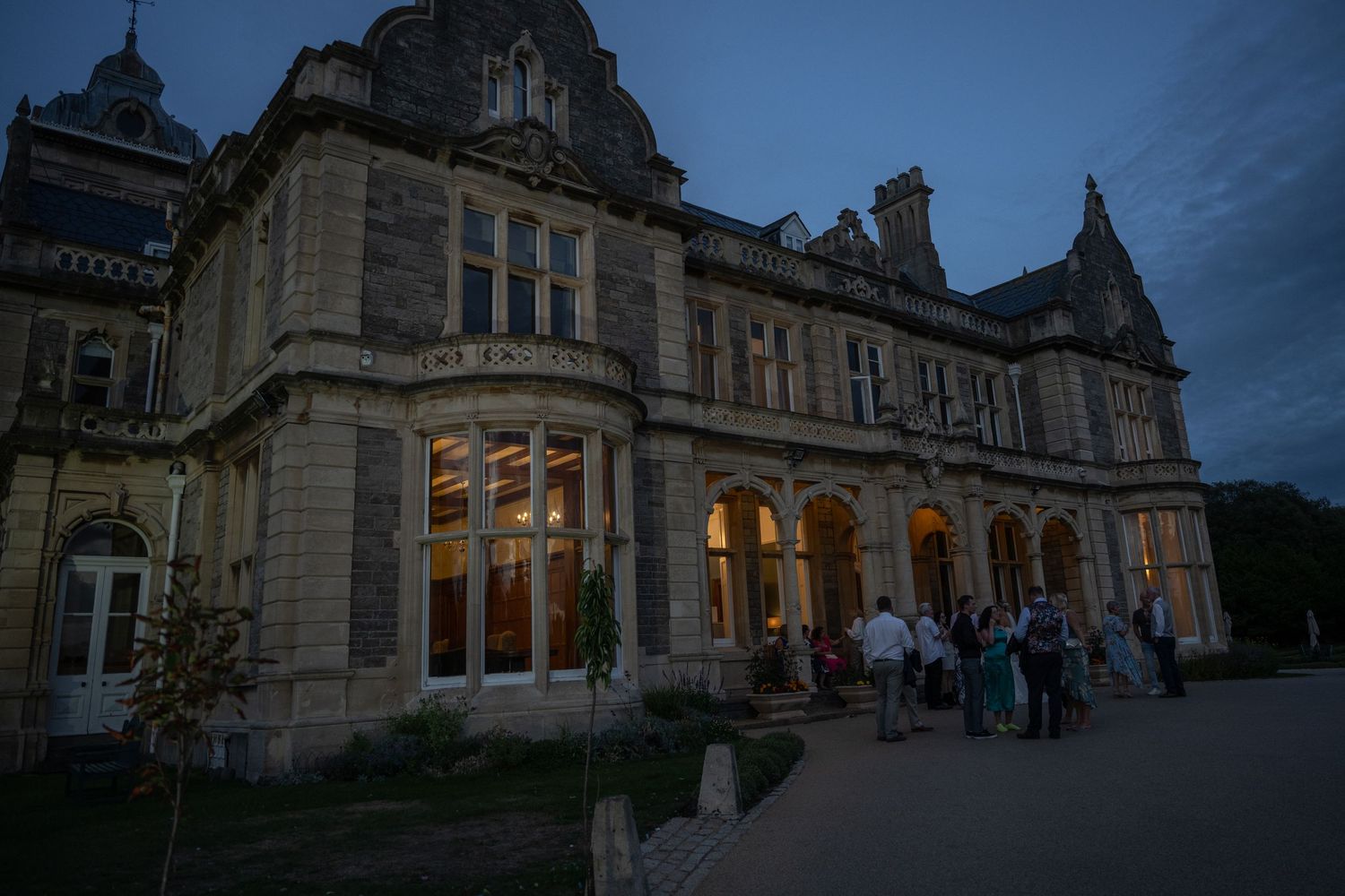 Historic stone mansion at dusk with illuminated windows and people gathered outside.