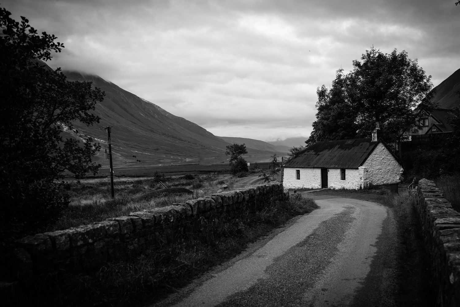 Black and white photograph of a lonely white cottage on a rural Scottish road with mountains behind.