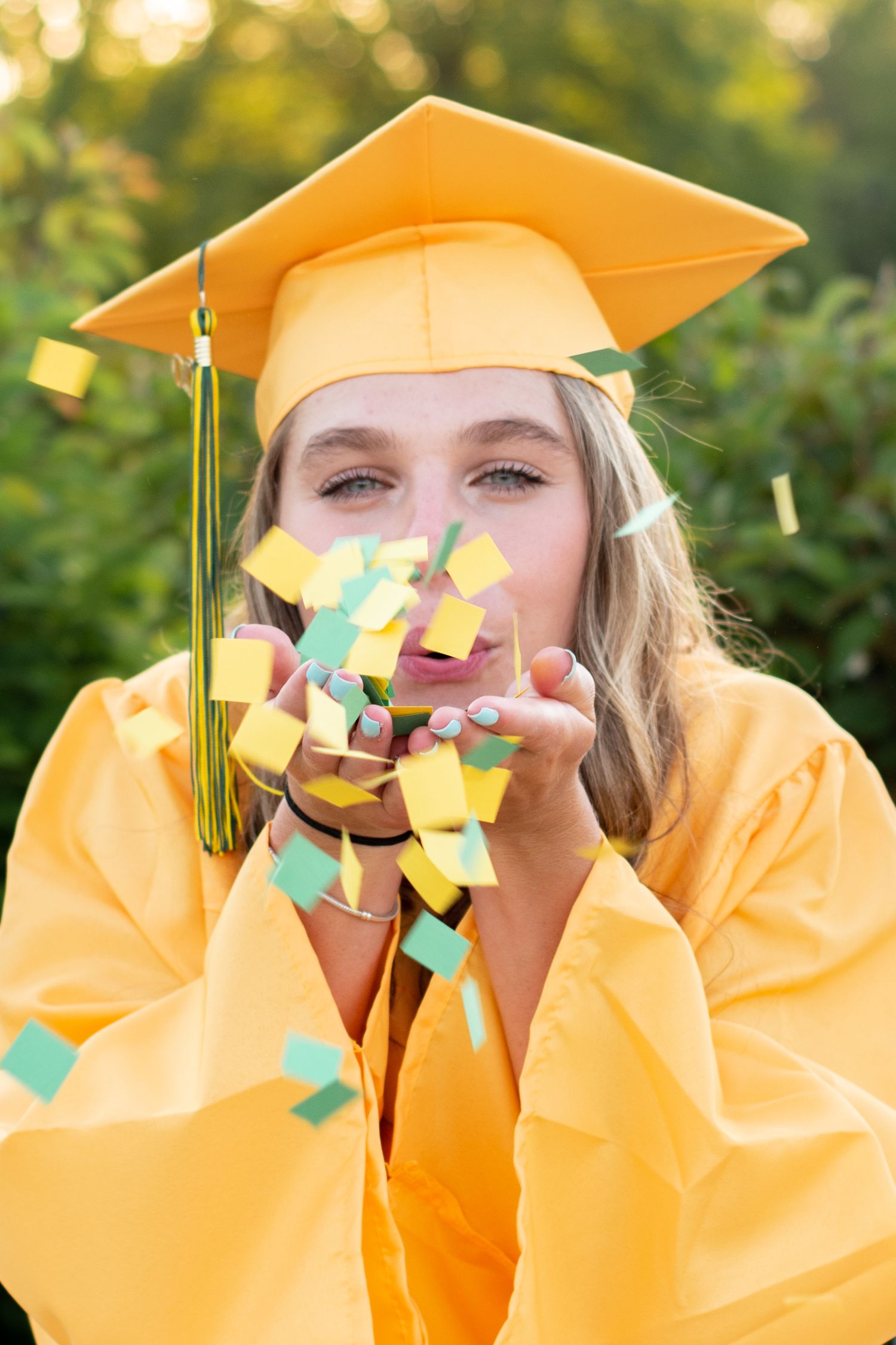 high school senior girl in gold cap and gown blowing confetti at sunset