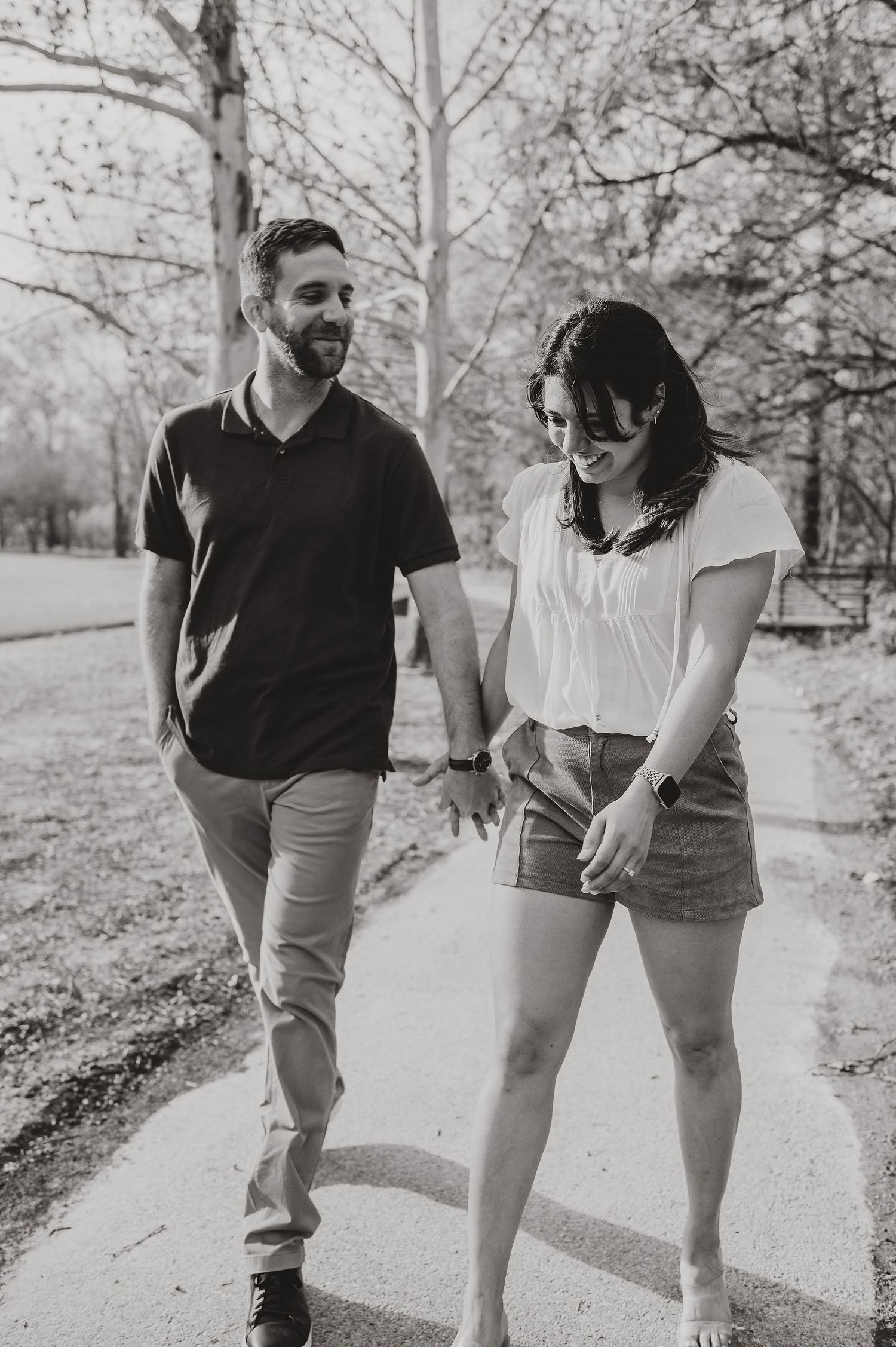A romantic couple walks together and laughs on a tree-lined path in black and white.
