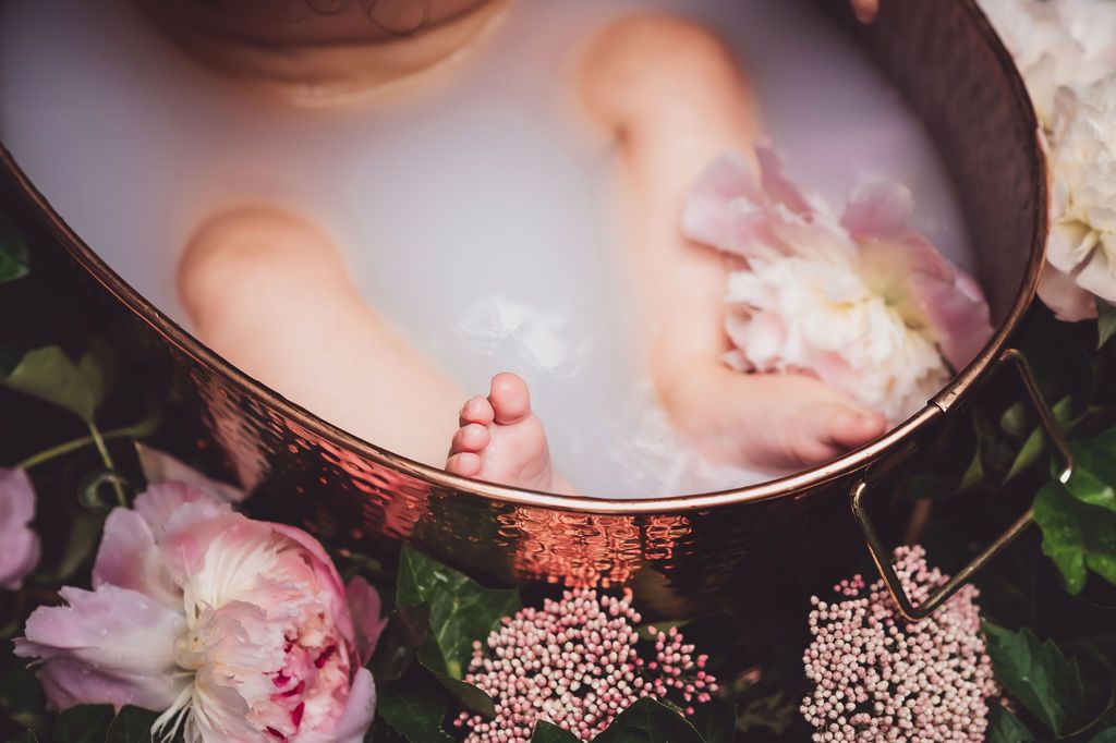 just the legs and toes of a baby are visible in a milk bath with peony flowers in and around a copper tub set in ivy