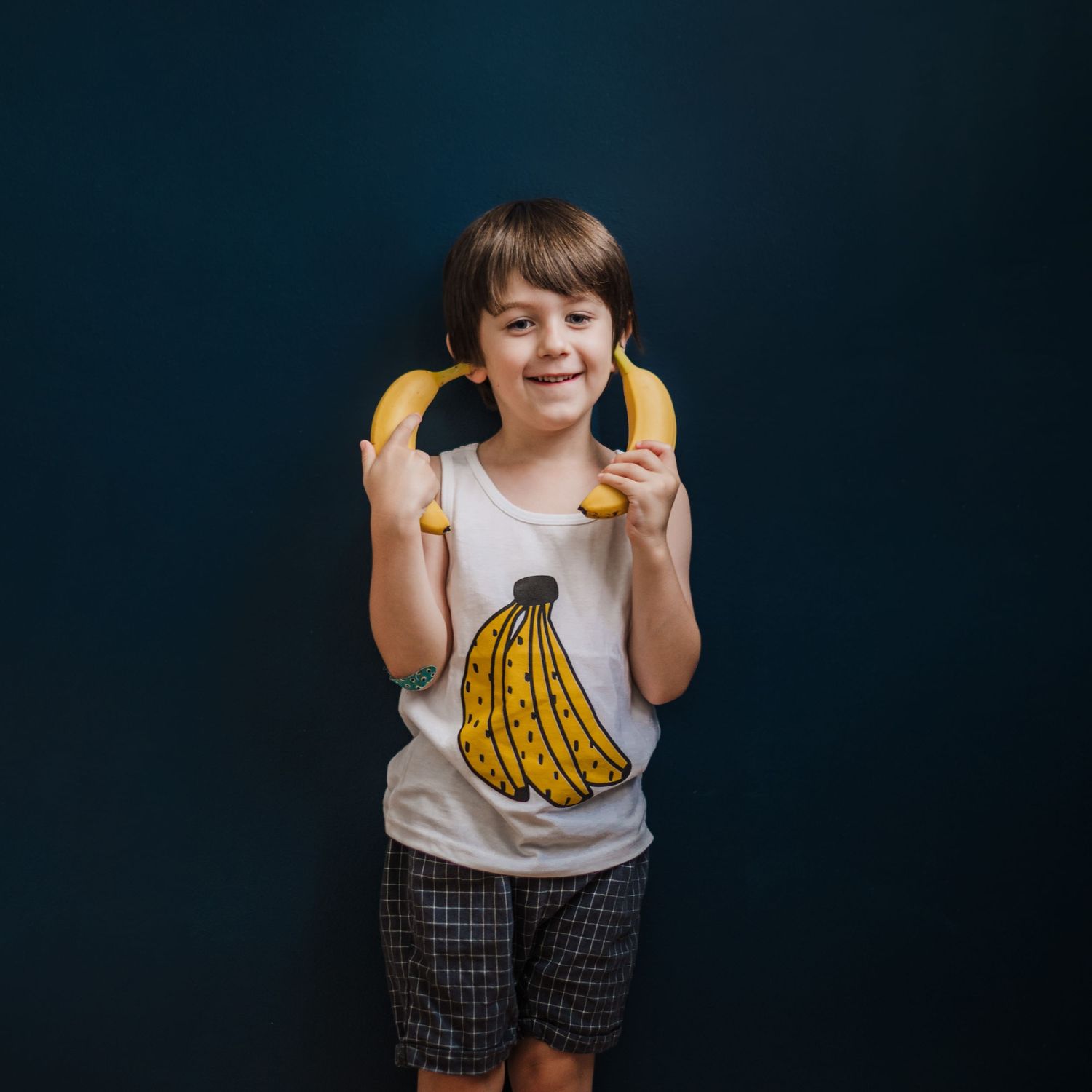 Child wearing banana graphic tank top holds fresh bananas as phones against dark background while smiling.