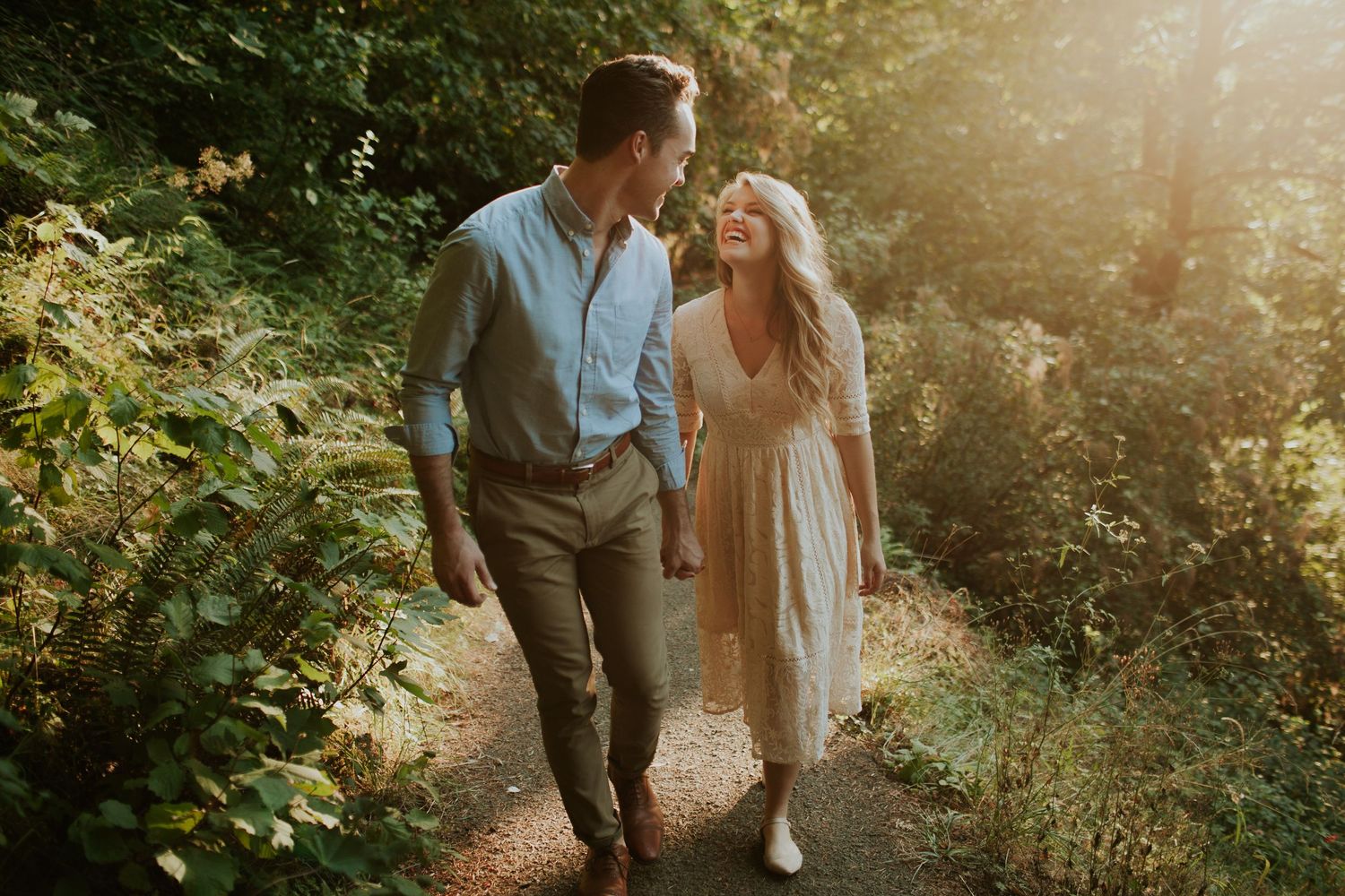 A couple walks hand in hand along a sunlit forest path during their engagement session.