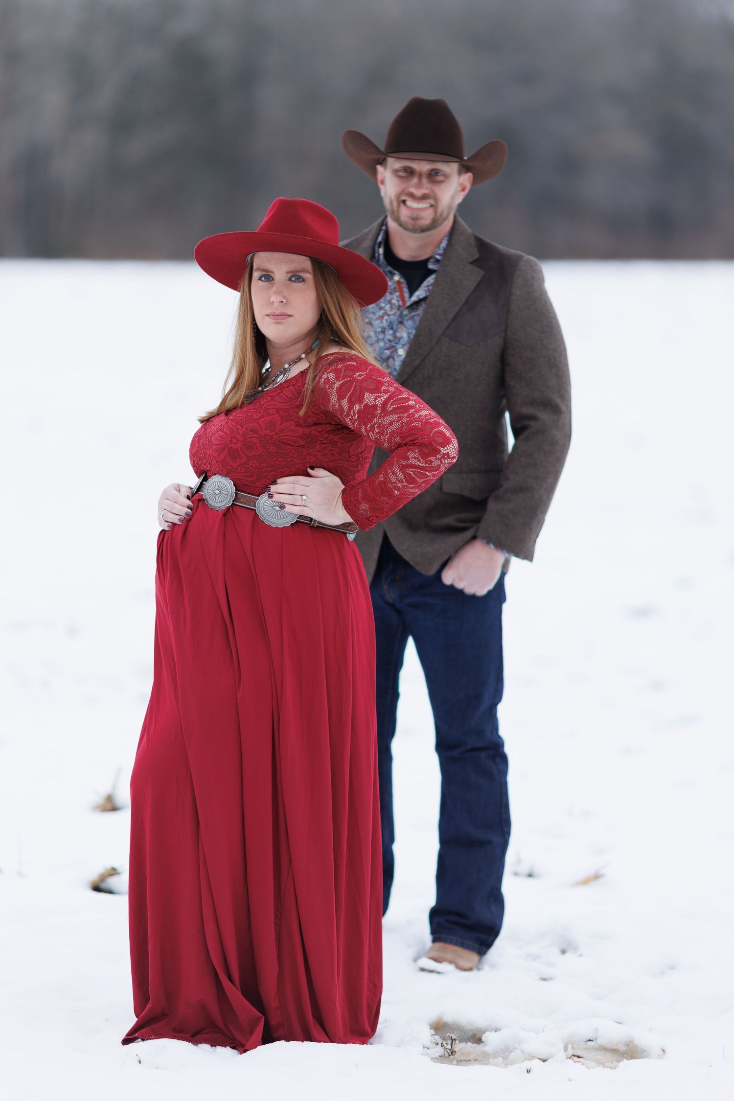 A pregnant cowgirl showing off her bump while her husband stands proudly in the background.