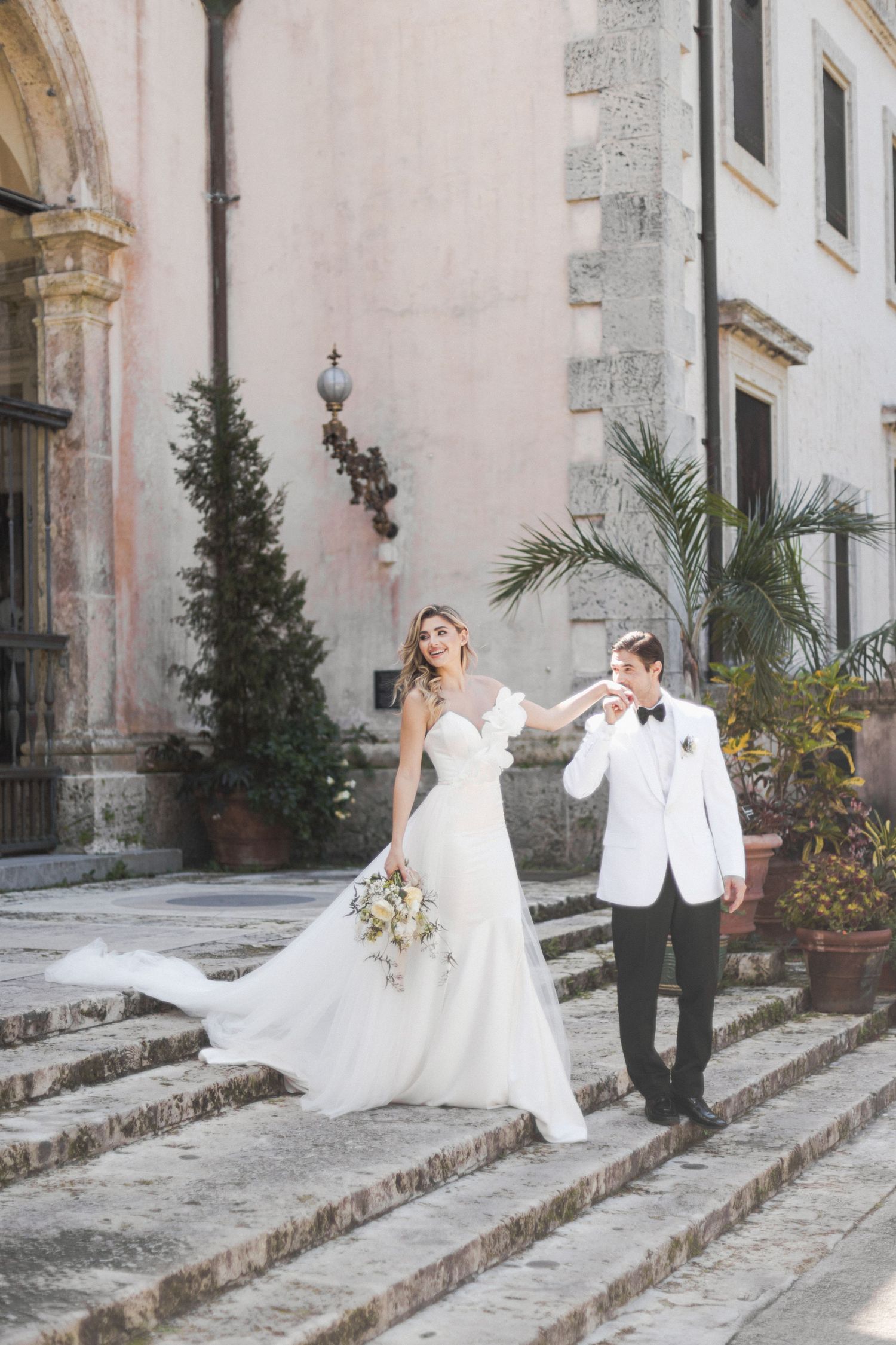 A romantic wedding portrait on stone steps with classic architecture and tropical plants.