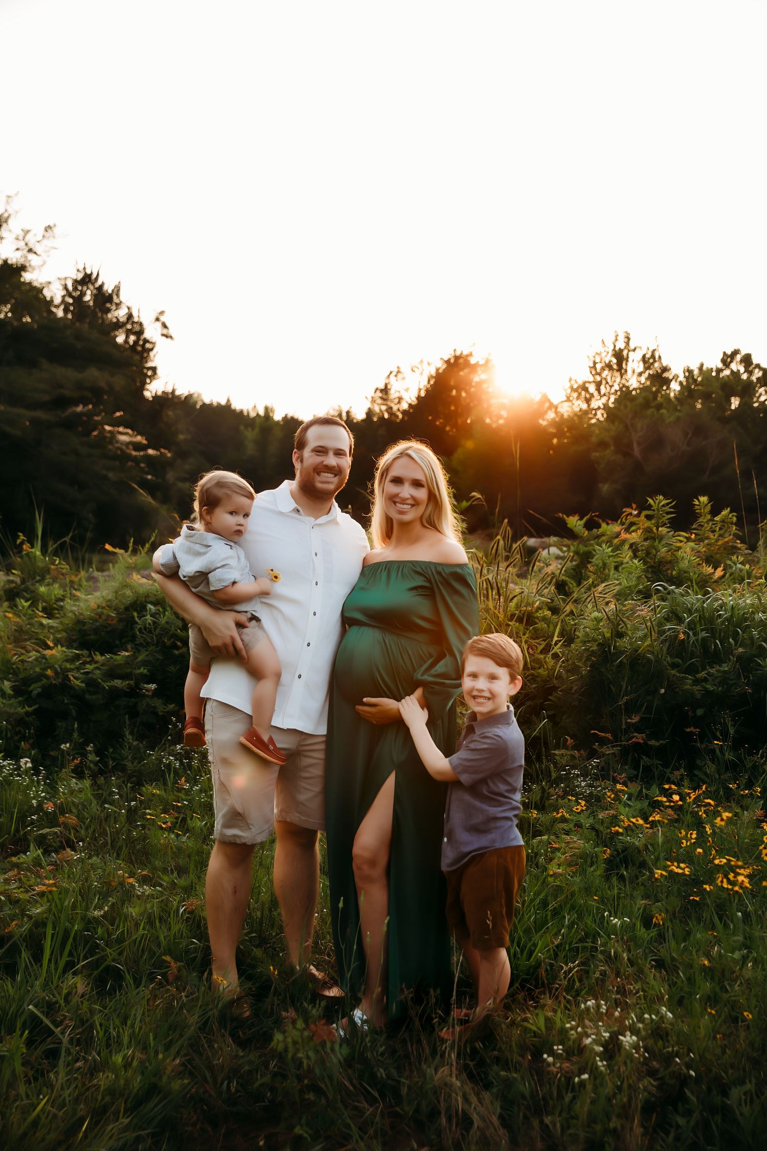 Family standing in wildflower field at sunset with expecting mother in green dress.