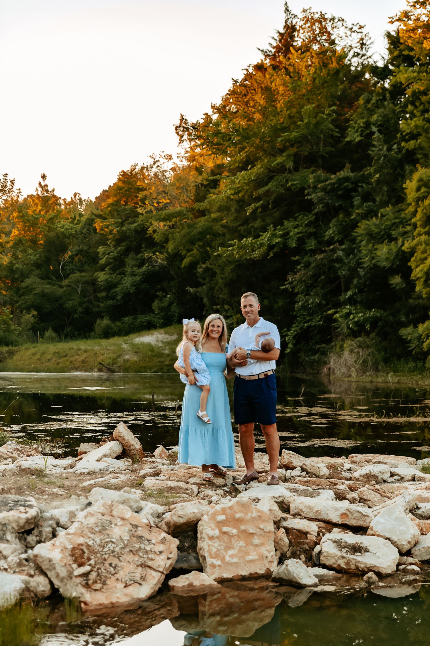 Family standing on rocky riverbank during golden hour with forest backdrop.