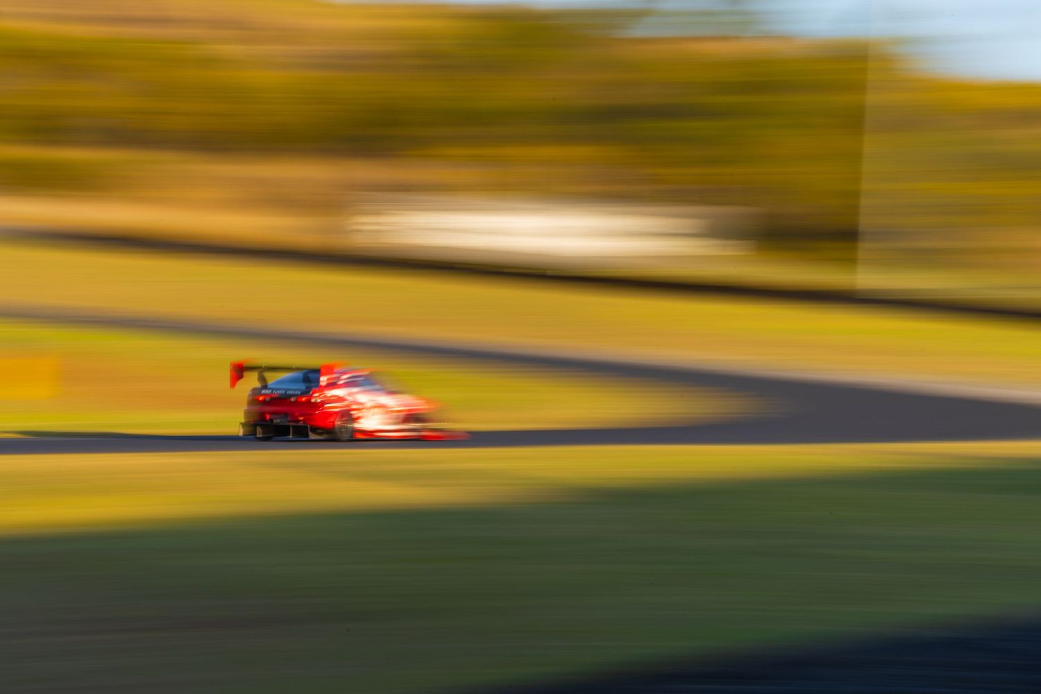 Motion-blurred red racecar speeding around a sunny racetrack bend.