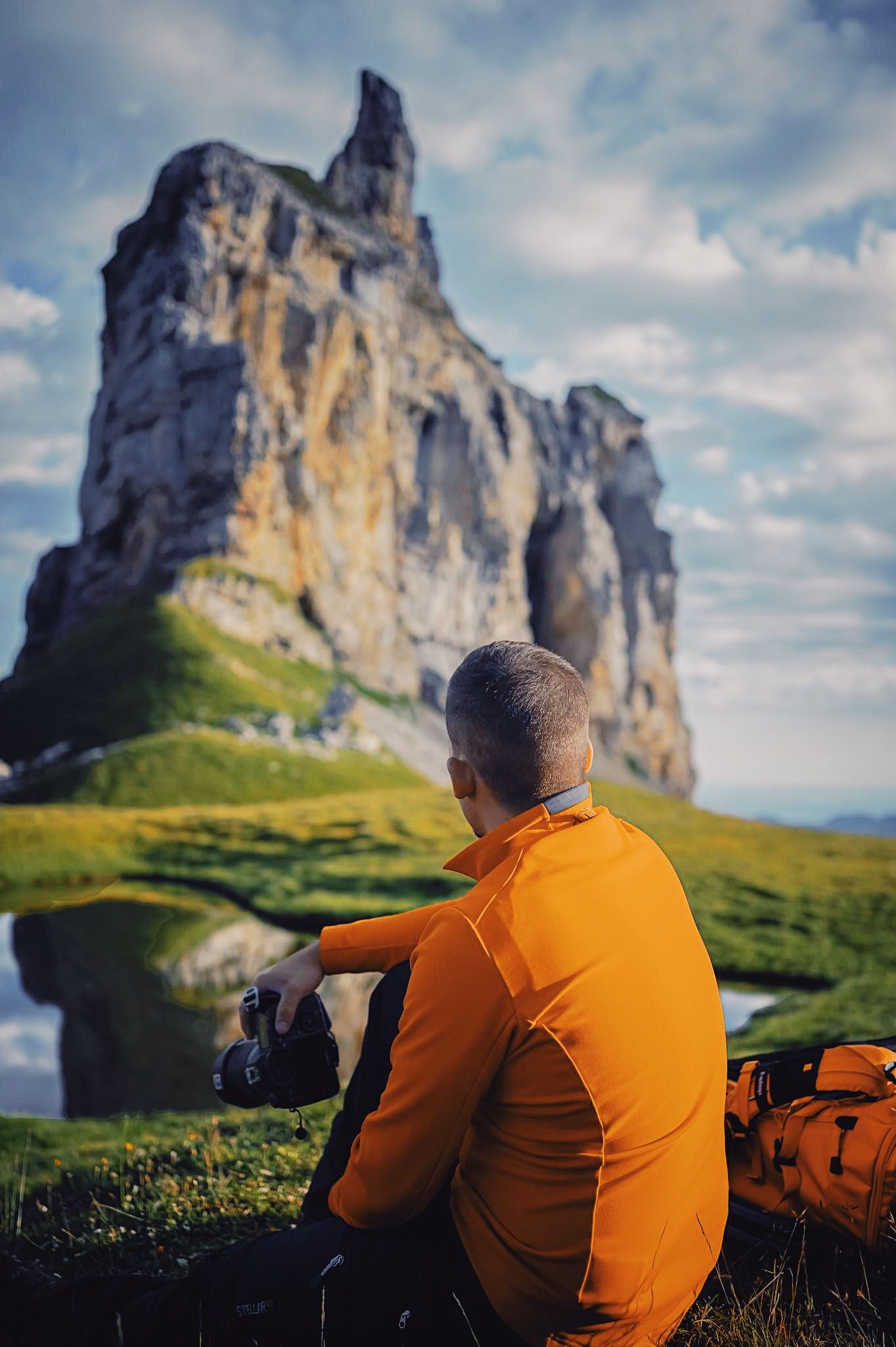 Photographer in orange shirt resting near dramatic mountain peak in scenic landscape.