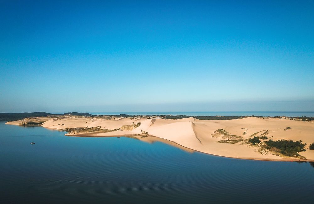 Aerial view of sand dunes meeting turquoise water under bright blue sky.
