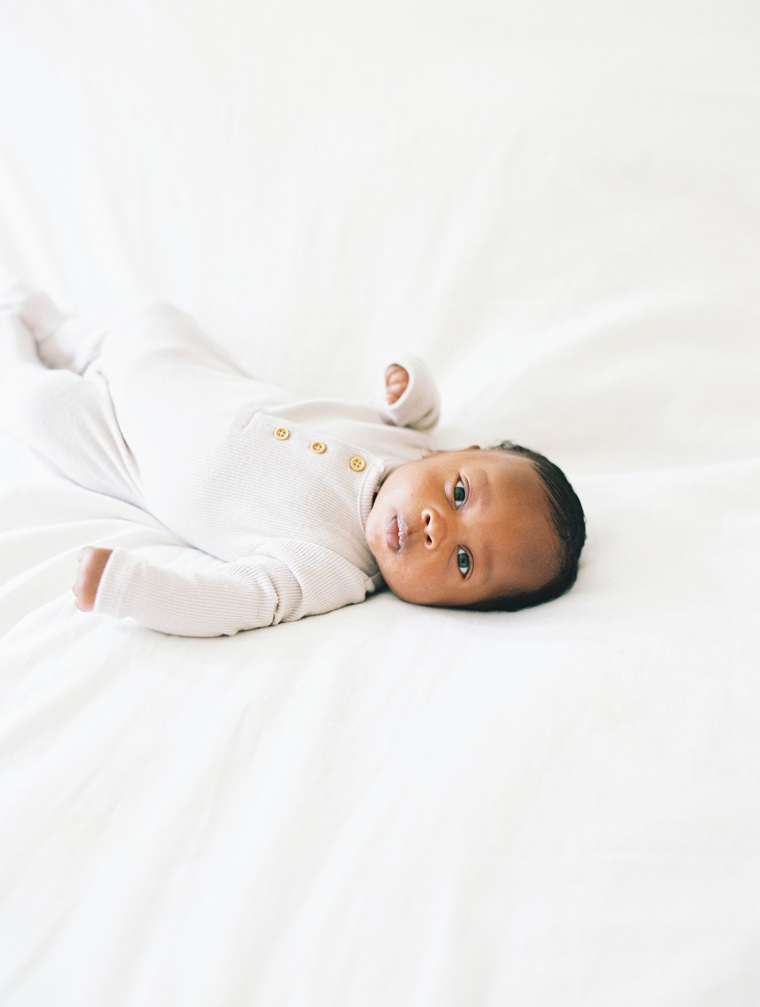Baby in white onesie lying on white bedding looking at camera.