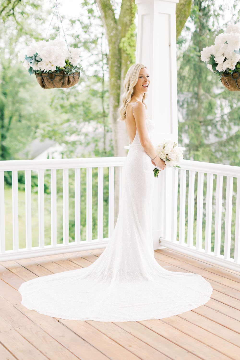 An elegant wedding dress with long train on white porch with hanging floral baskets.