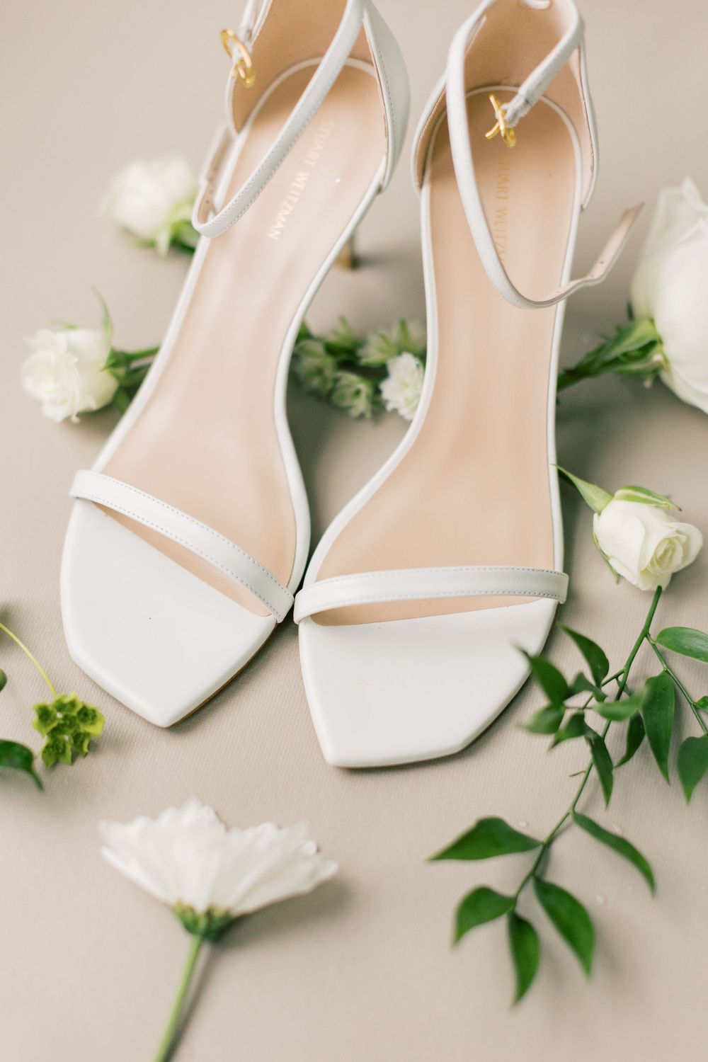 Elegant white bridal shoes styled with fresh white roses and greenery on beige background.