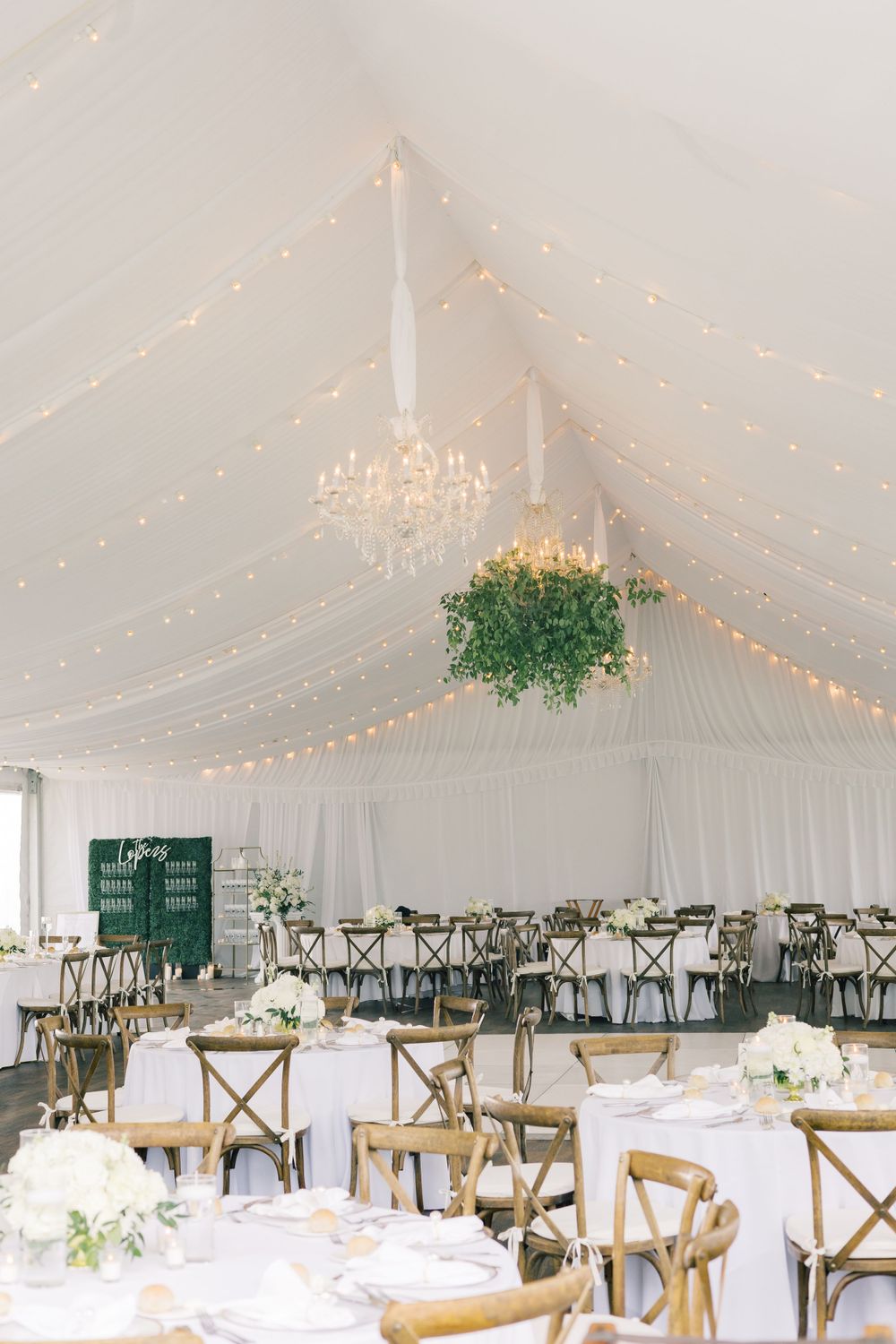 White wedding tent interior with string lights, chandelier and cross-back chairs.