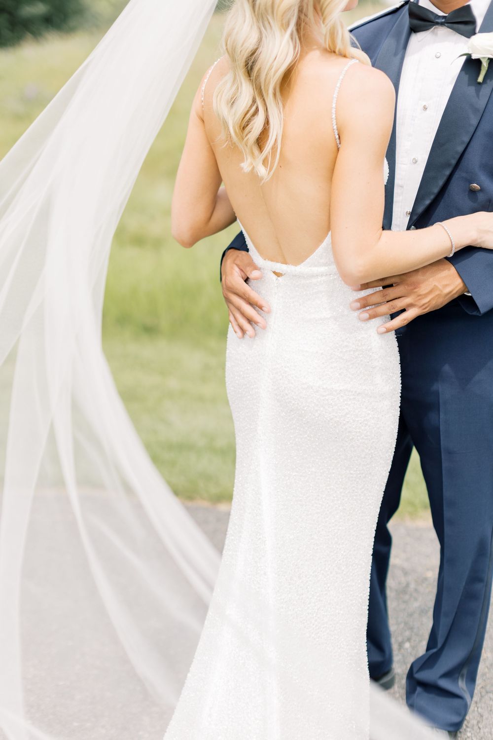 Elegant backless white wedding dress with flowing veil and navy tuxedo shown in romantic outdoor setting.