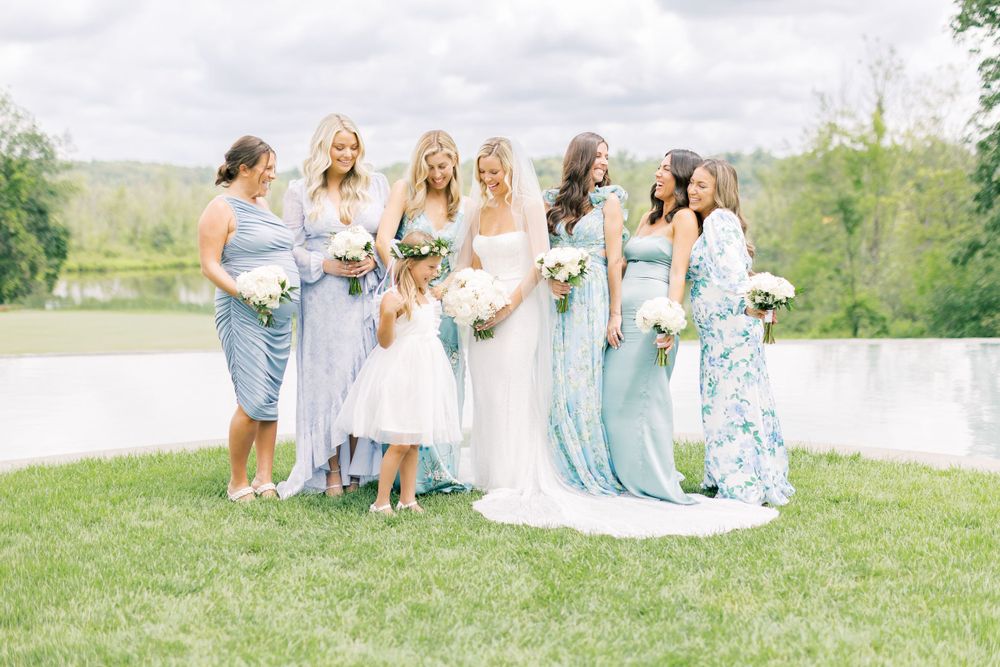 A bridal party in soft blue dresses poses together on a lakeside lawn.