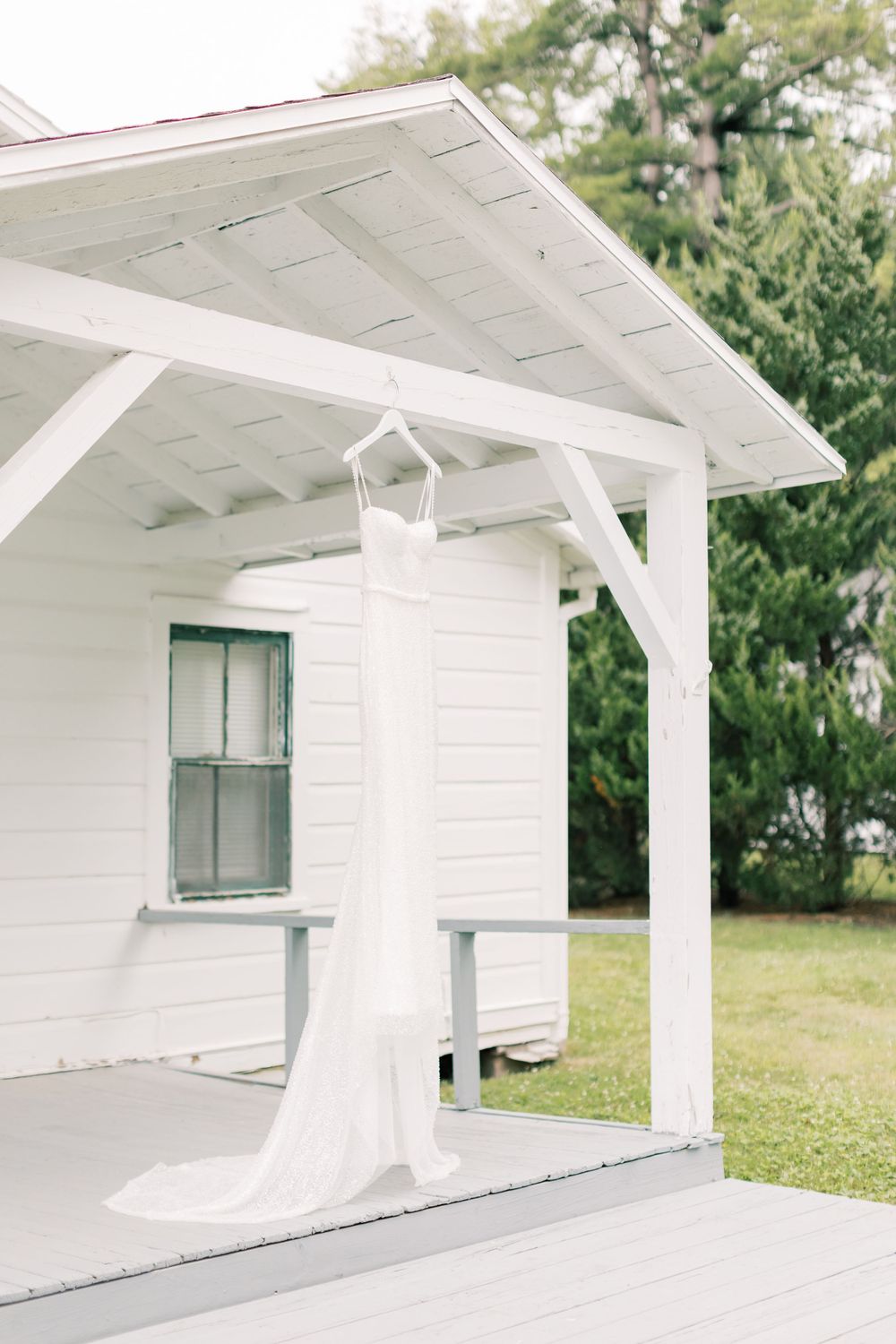 A white wedding dress hangs elegantly from a white porch roof of a charming cottage.