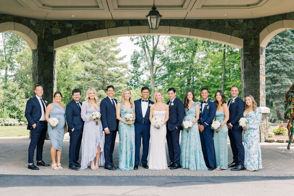 Wedding party in formal attire poses under stone archway surrounded by greenery.