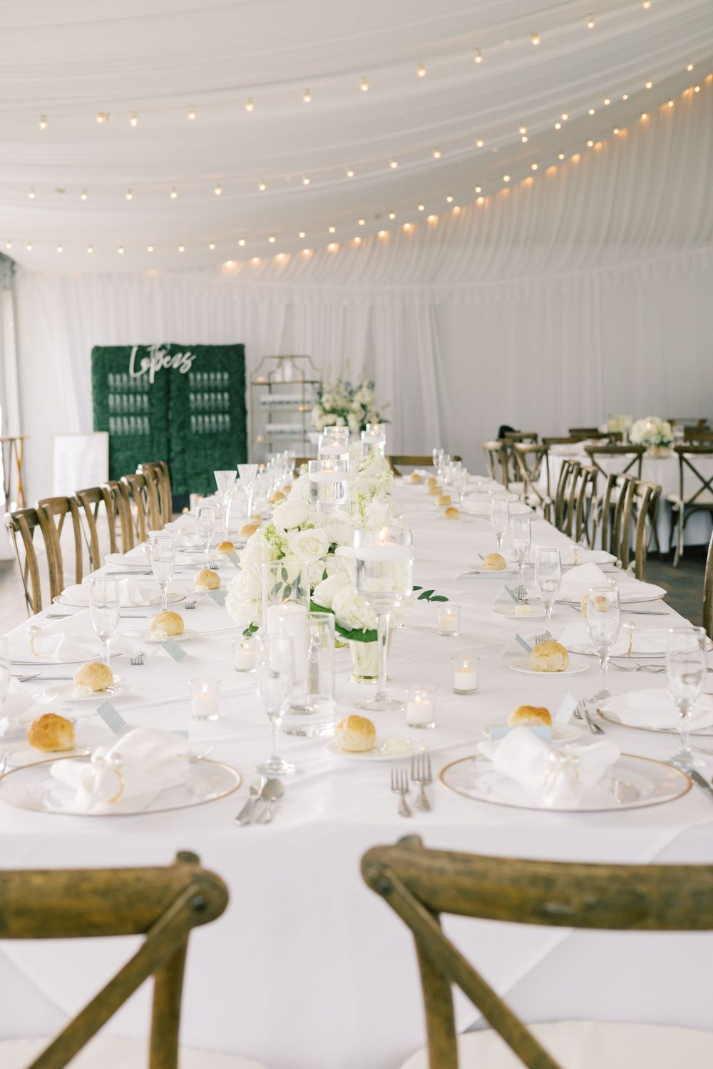 Elegant white wedding reception tent with long tables, string lights, and delicate place settings.