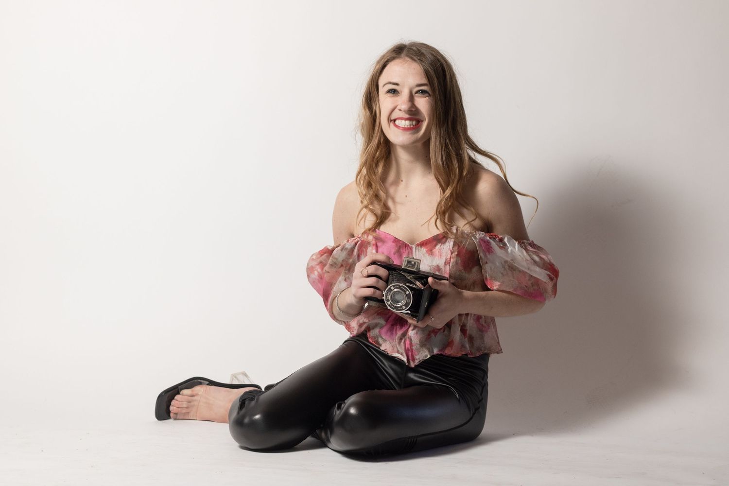 Professional photographer sitting on floor with vintage camera in pink off-shoulder top.