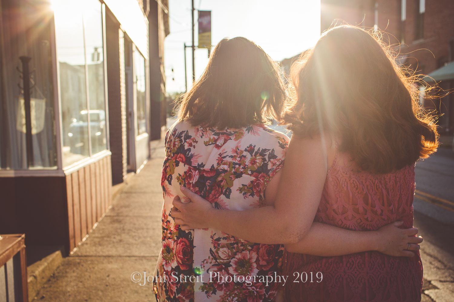 Friends in floral and lace tops embrace while walking down a sunlit street.