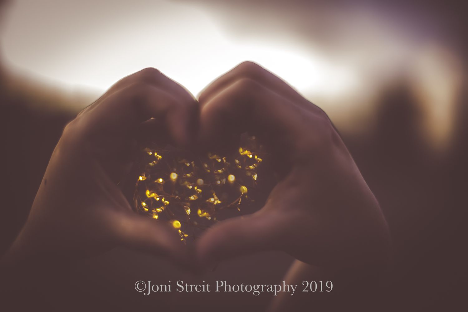 Hands forming a heart shape against sunset with golden bokeh lights shining through.