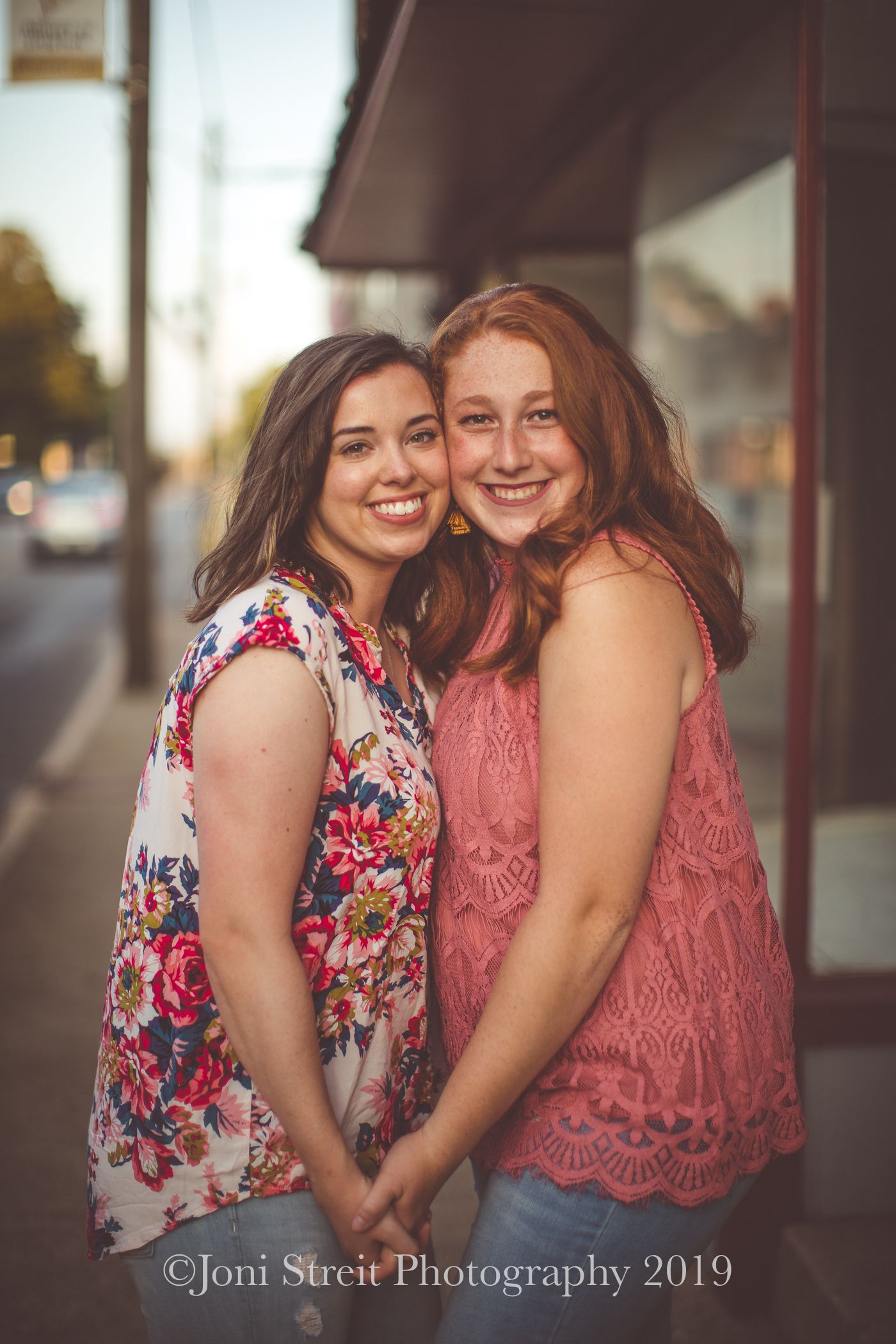Two friends in floral and pink lace tops share a candid moment on a city sidewalk at sunset.