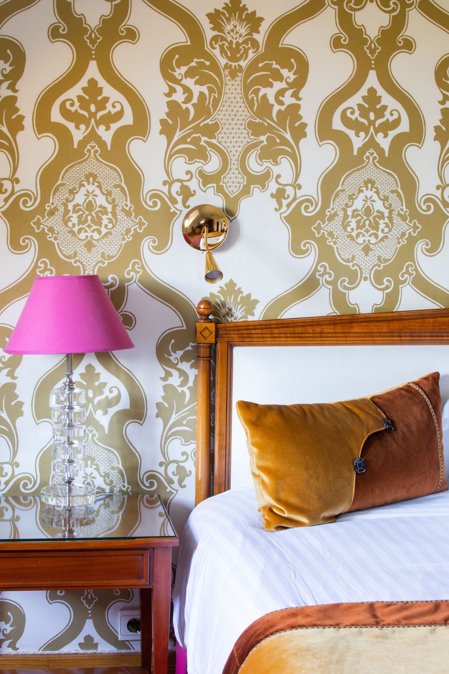 Bedroom with ornate gold damask wallpaper, pink lamp, and velvet pillows on bed.