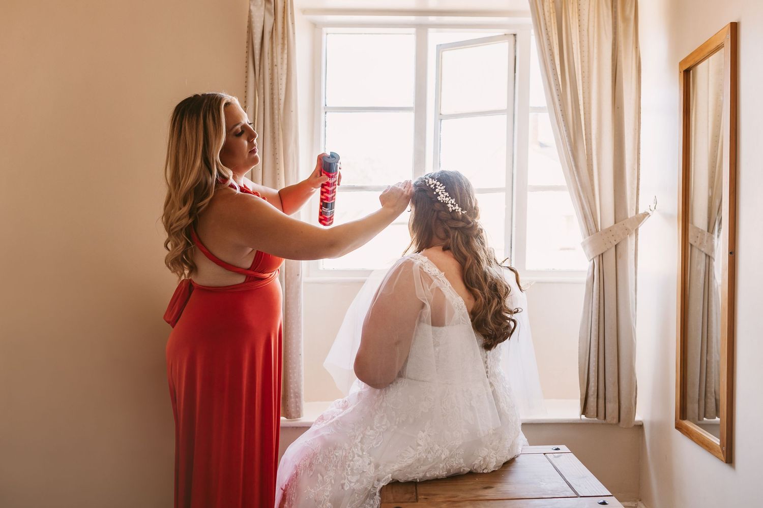 Friend spraying Brides hair with hairspray in front of bright window
