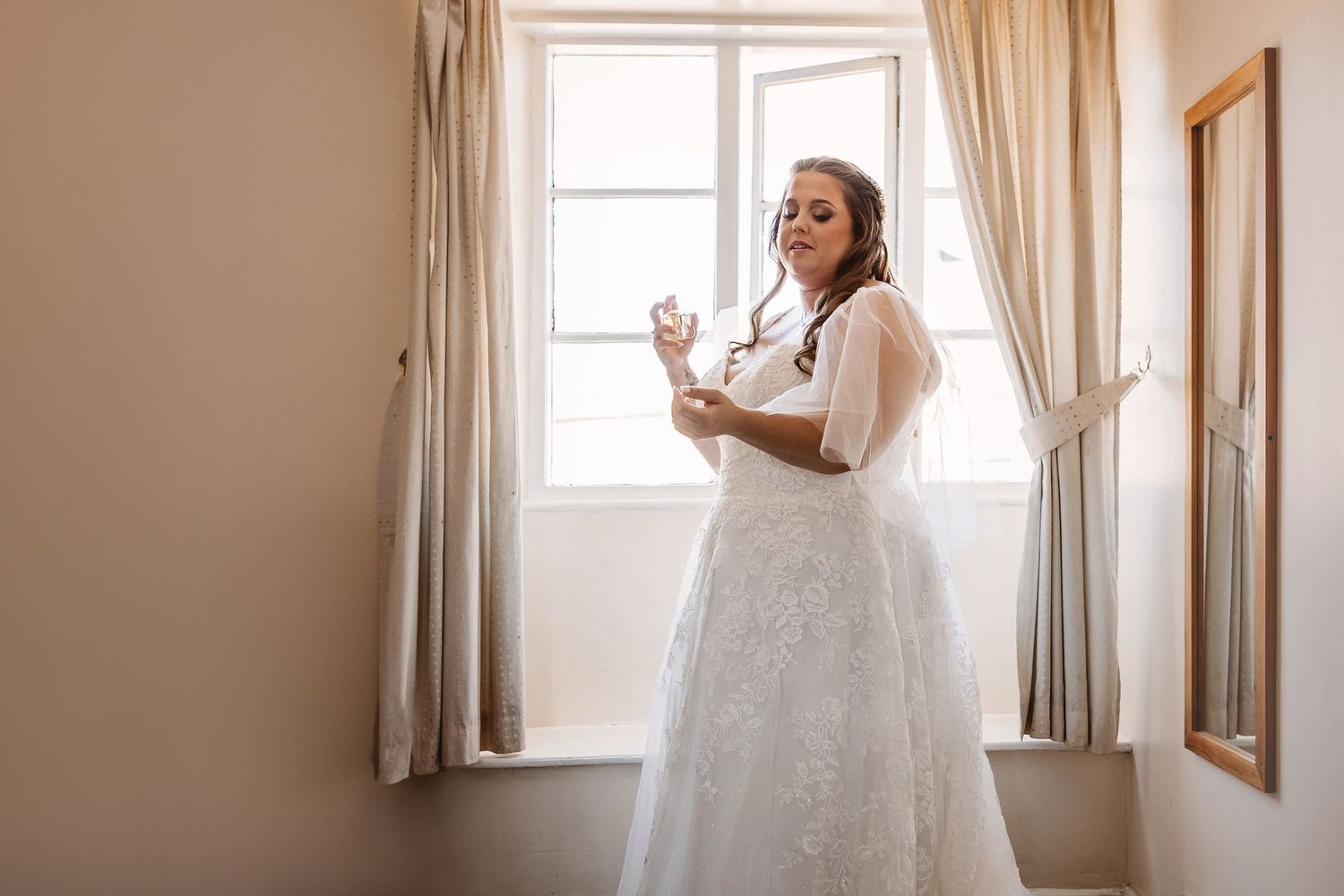 Bride standing in front of bright window spraying perfume on herself